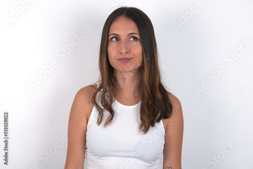 young beautiful caucasian woman wearing white top over white background has worried face looking up lips together, being upset thinking about something important, keeps hands down.