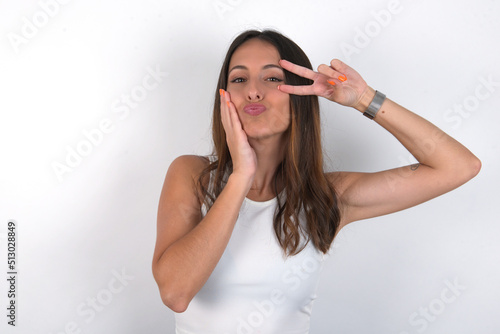 young beautiful caucasian woman wearing white top over white background  making v-sign near eyes. Leisure lifestyle people person celebrate flirt coquettish concept. © Jihan