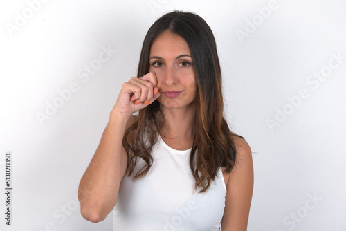 Unhappy young beautiful caucasian woman wearing white top over white background crying while posing at camera whipping tears with hand.