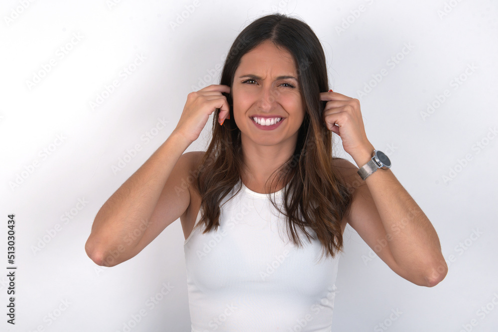 Happy young beautiful caucasian woman wearing white Top over white background ignores loud music and plugs ears with fingers asks to turn off sound Happy young beautiful caucasian woman wearing white Top over white background ignores loud music and plugs ears with fingers asks to turn off sound