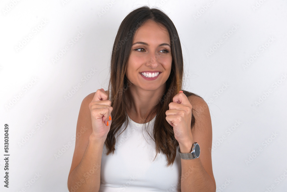 young beautiful caucasian woman wearing white Top over white background clenches fists and awaits for something nice happened looks away bites lips and waits announcement of results young beautiful caucasian woman wearing white Top over white background clenches fists and awaits for something nice happened looks away bites lips and waits announcement of results