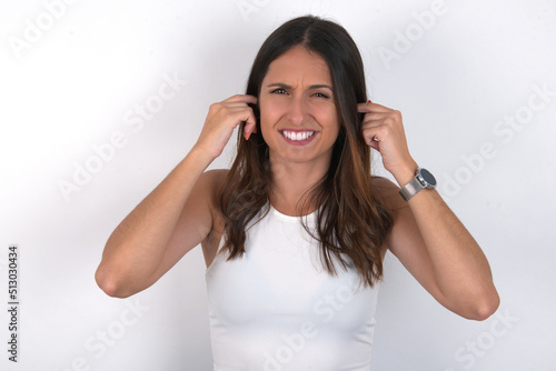 Happy young beautiful caucasian woman wearing white Top over white background ignores loud music and plugs ears with fingers asks to turn off sound
