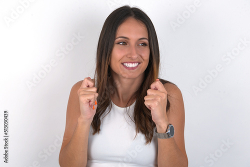 young beautiful caucasian woman wearing white Top over white background clenches fists and awaits for something nice happened looks away bites lips and waits announcement of results