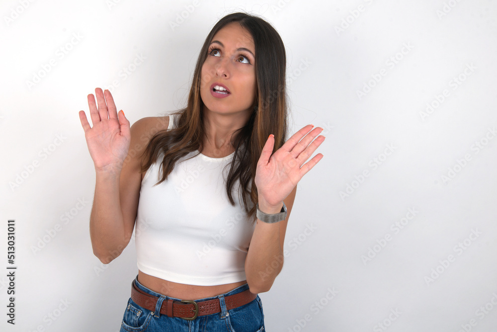 young beautiful caucasian woman wearing white Top over white background keeps palms forward and looks with fright above on ceiling tries to defense herself from invisible danger opens mouth. young beautiful caucasian woman wearing white Top over white background keeps palms forward and looks with fright above on ceiling tries to defense herself from invisible danger opens mouth.
