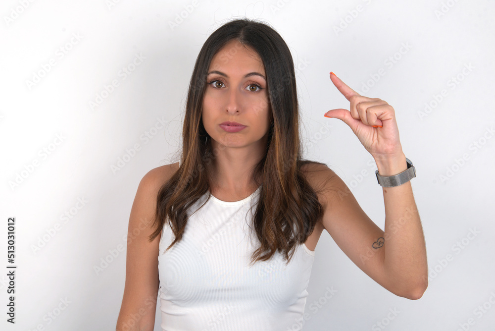 young beautiful caucasian woman wearing white Top over white background purses lip and gestures with hand, shows something very little. young beautiful caucasian woman wearing white Top over white background purses lip and gestures with hand, shows something very little.
