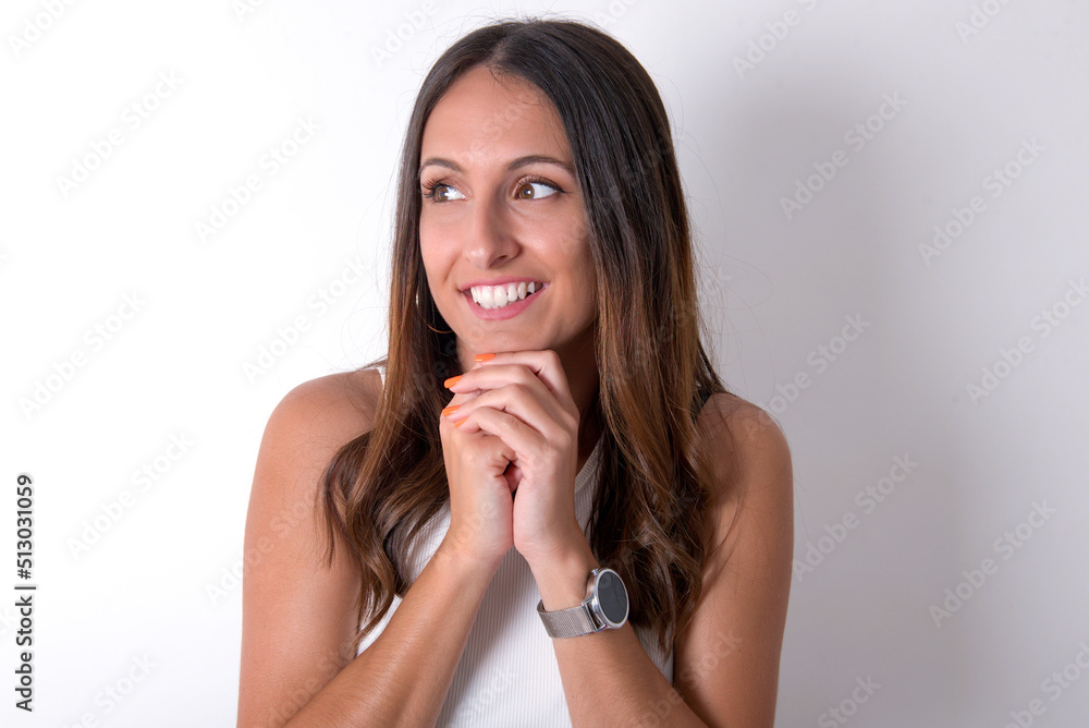 Happy young beautiful caucasian woman wearing white Top over white background, anticipates something awesome happen, looks happily aside, keeps hands together near face, has glad expression. Happy young beautiful caucasian woman wearing white Top over white background, anticipates something awesome happen, looks happily aside, keeps hands together near face, has glad expression.