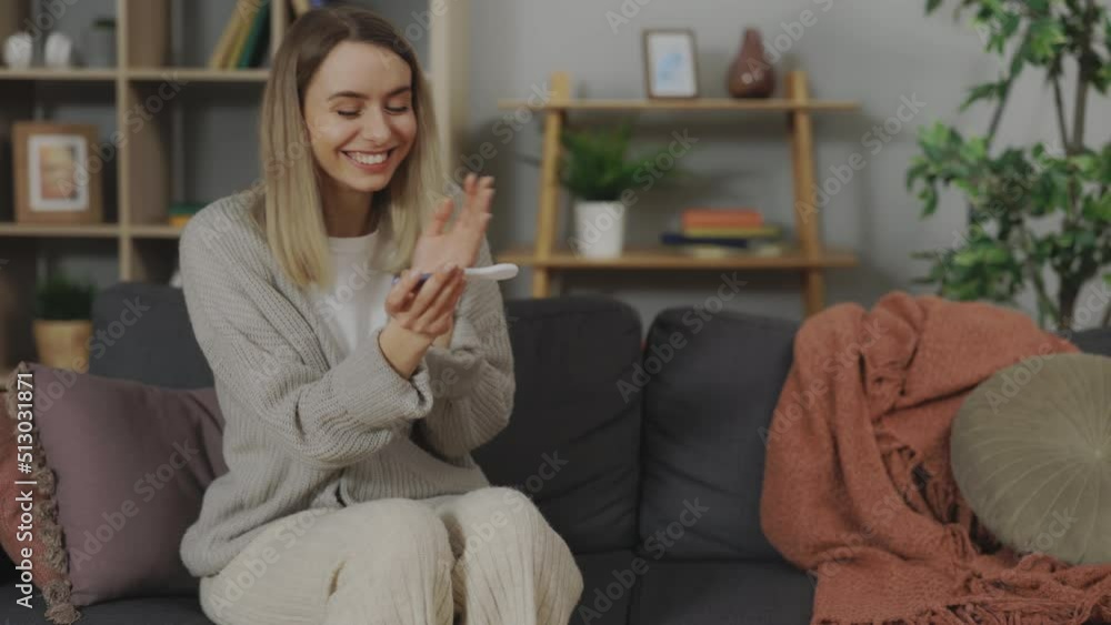 Caucasian woman with blond hair checking pregnancy test while sitting on couch. Cheerful young female in casual clothes celebrating great news at home.