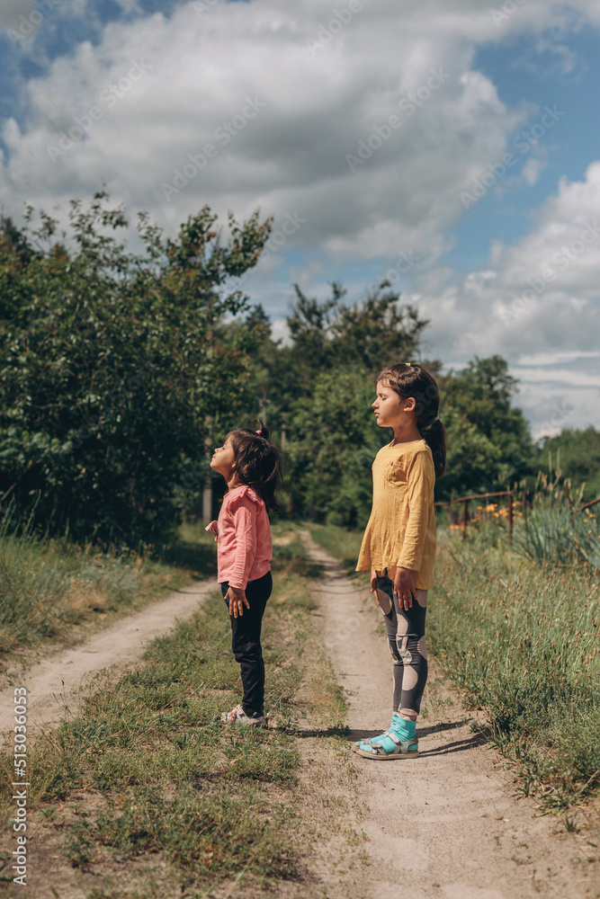 Two little girls with closed eyes raised their heads up to the sky with faith and hope for a better future, Ukrainian children Two little girls with closed eyes raised their heads up to the sky with faith and hope for a better future, Ukrainian children