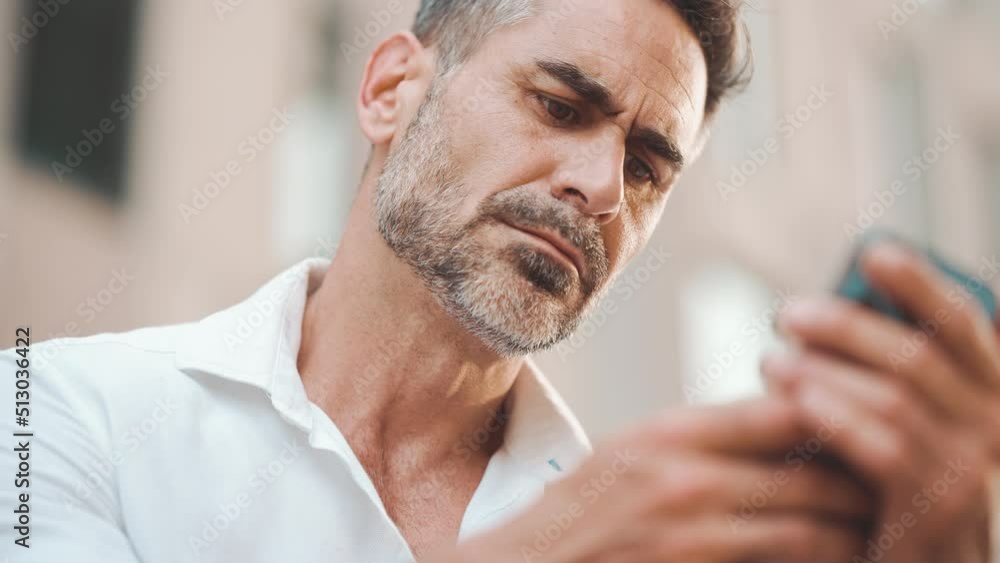 Clouse-up, mature businessman with neat beard uses mobile phone sits on bench in the financial district in the city. Man scrolls through information on smartphone. Camera zoom Clouse-up, mature businessman with neat beard uses mobile phone sits on bench in the financial district in the city. Man scrolls through information on smartphone. Camera zoom