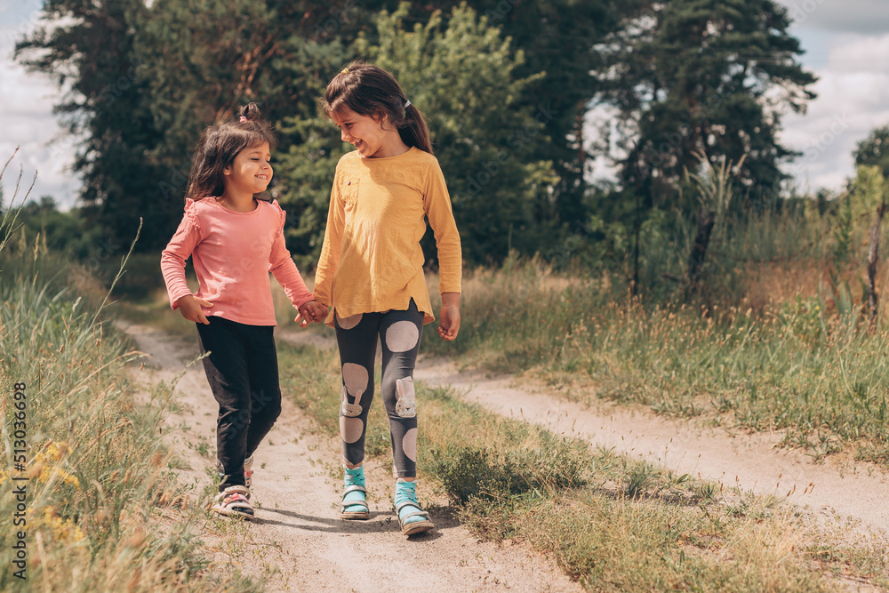 Little girl friends walk holding hands along a rural path near the forest, summer vacation