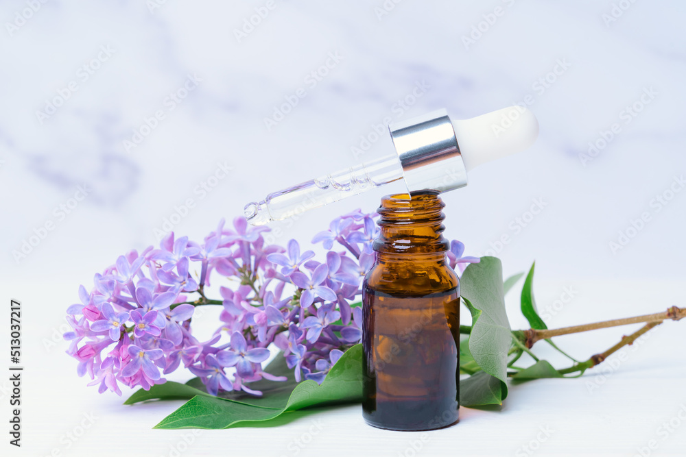 A bottle of lilac essential oil and a branch with flowers on a white marble background A bottle of lilac essential oil and a branch with flowers on a white marble background