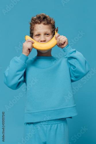  handsome, happy boy stands in blue clothes on a blue background and holds a banana in his hand, substituting it as a smile to his face. Studio photo with empty space for advertising insert