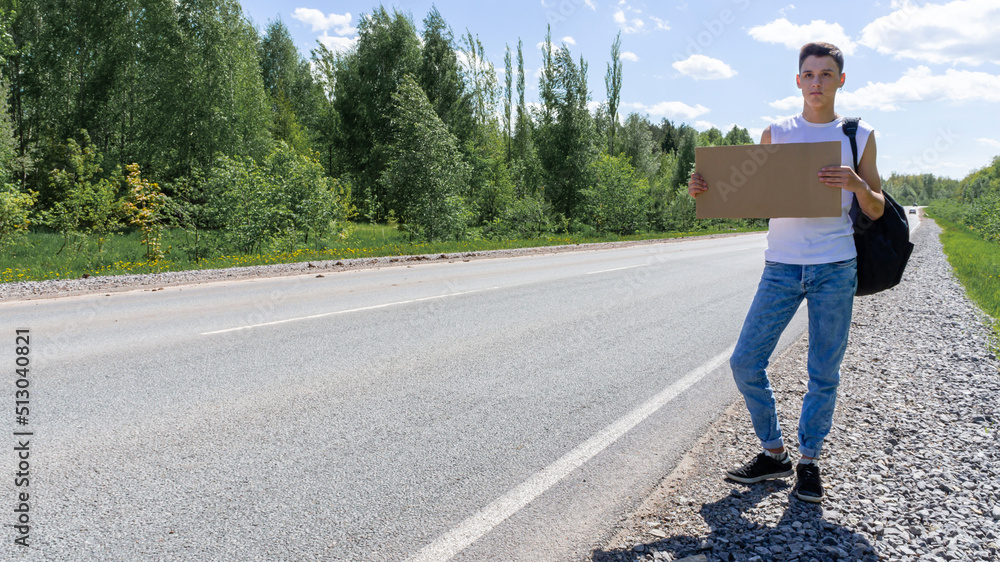 A young man hitchhiking is standing by the road with an empty cardboard box. The concept of travel, adventure, outdoor activities, vacations. High quality photo