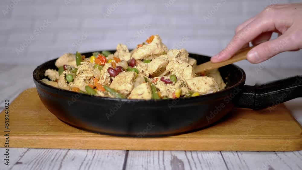 Female Hand Mixing Fried Chicken Meat with Vegetables in a Frying Pan. Bright pieces of steamed fillet mixed with colorful vegetables. Green peas, beans, corn, carrots. Cutting board. Wooden spatula. Female Hand Mixing Fried Chicken Meat with Vegetables in a Frying Pan. Bright pieces of steamed fillet mixed with colorful vegetables. Green peas, beans, corn, carrots. Cutting board. Wooden spatula.
