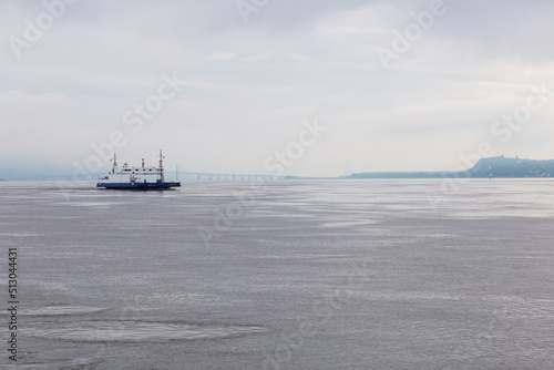 Ferry boat moving on the St. Lawrence River towards the Lévis port during a hazy summer morning, with the Island of Orleans bridge in soft focus background, Quebec City, Quebec, Canada