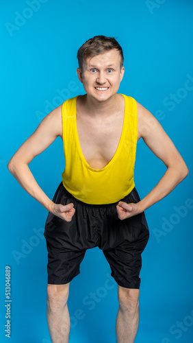 thin athlete in a yellow t-shirt and black shorts on a blue background. funny shakes muscles, goes in for sports, fitness
