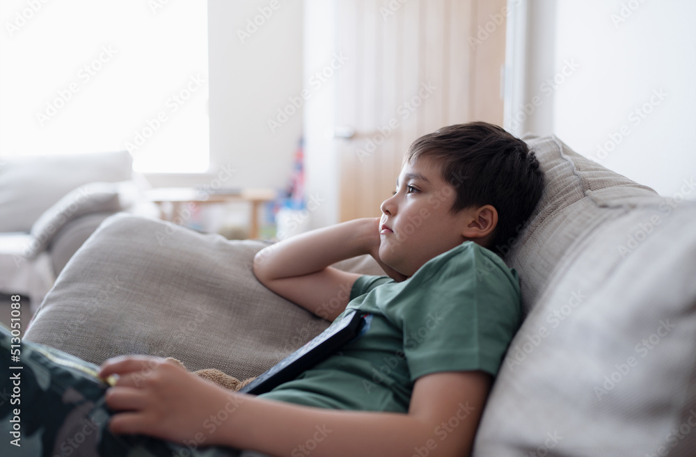 Kid with remote control and looking up with curious face,Young boy sitting on sofa watching cartoon on TV, Portrait Child lying down on couch relaxing in living room after back from school. Kid with remote control and looking up with curious face,Young boy sitting on sofa watching cartoon on TV, Portrait Child lying down on couch relaxing in living room after back from school.