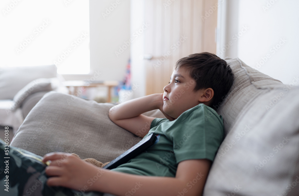 Kid with remote control and looking up with curious face,Young boy sitting on sofa watching cartoon on TV, Portrait Child lying down on couch relaxing in living room after back from school.