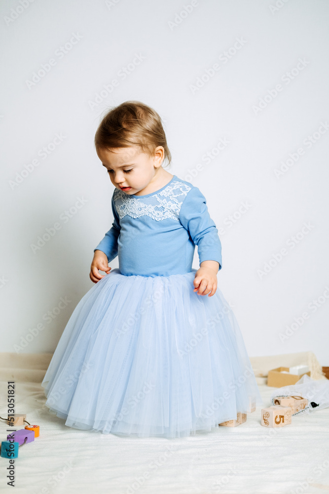 Cute baby girl in blue princess dress playing with toys at home. Kids dresses for prom and birthday holiday Cute baby girl in blue princess dress playing with toys at home. Kids dresses for prom and birthday holiday