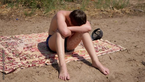 Bored Child Sitting on a Coverlet on the Sand, Sunbathing on Beach in Hot Sun. Tired offended boy with his head down, resting in nature in the bright scorching sun. Heat, holidays, rest, summer.
