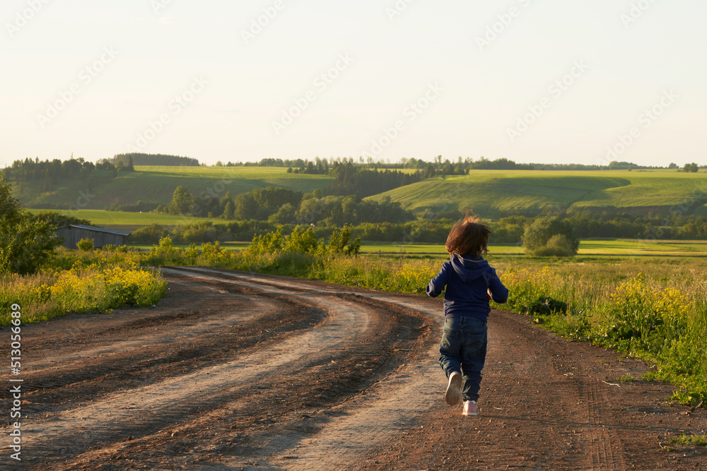 The child runs away into the distance along a dirt winding road passing through a green pasture. The soft light of the setting sun.
