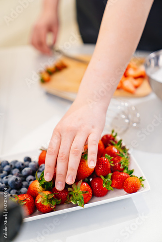 women s hand takes strawberries and blueberries from plate to decorate desserts.