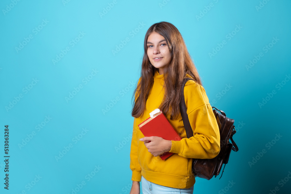 A young student with a backpack looks at a book and reads. Education and training, visiting the library. Young woman in yellow hoodie and jeans on a blue background, copy space A young student with a backpack looks at a book and reads. Education and training, visiting the library. Young woman in yellow hoodie and jeans on a blue background, copy space