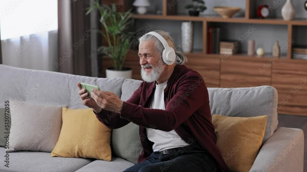 Happy handsome elderly gray-haired man in white headphones holding smartphone and laughing. Old man sitting on couch at home watching funny content videos blogs, TV shows, films. Happy handsome elderly gray-haired man in white headphones holding smartphone and laughing. Old man sitting on couch at home watching funny content videos blogs, TV shows, films.