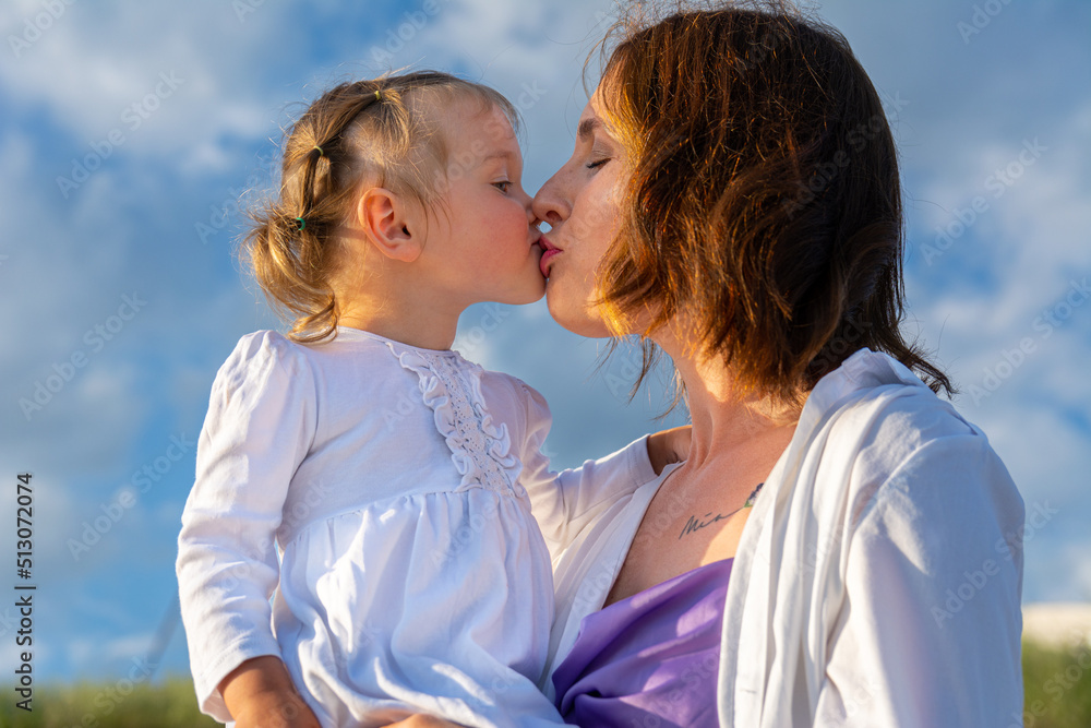 Portrait of a little girl and her mother. Mom and daughter are cute hugging against the blue sky with clouds. Mom and daughter in white clothes, family happiness. Portrait of a little girl and her mother. Mom and daughter are cute hugging against the blue sky with clouds. Mom and daughter in white clothes, family happiness.