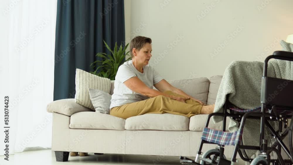 Senior woman massaging her legs while sitting on the couch near the wheelchair Senior woman massaging her legs while sitting on the couch near the wheelchair