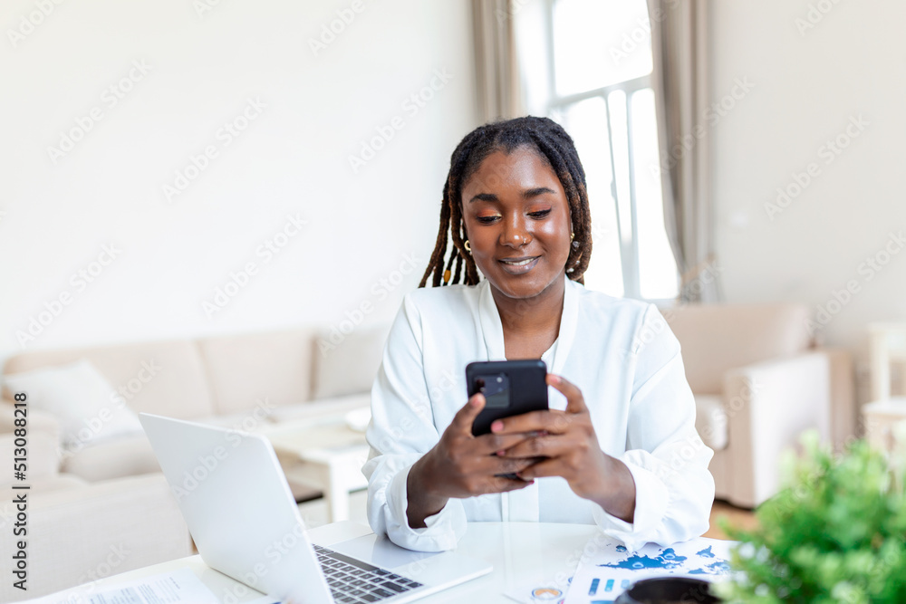 Young smiling African business woman using smartphone near computer in office Young smiling African business woman using smartphone near computer in office