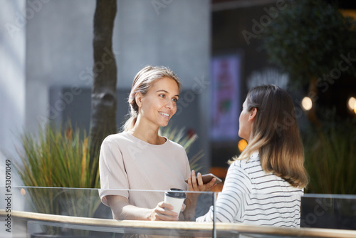 Positive young women in casual clothing standing at railing in contemporary lobby and sharing news from social medias