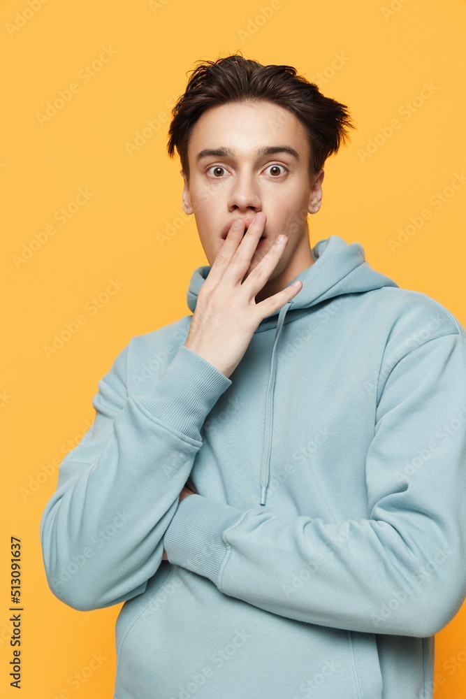 a close photo of a handsome young man standing on an orange background in a light blue hoodie with his arms crossed over his chest covering his mouth with his hand a close photo of a handsome young man standing on an orange background in a light blue hoodie with his arms crossed over his chest covering his mouth with his hand