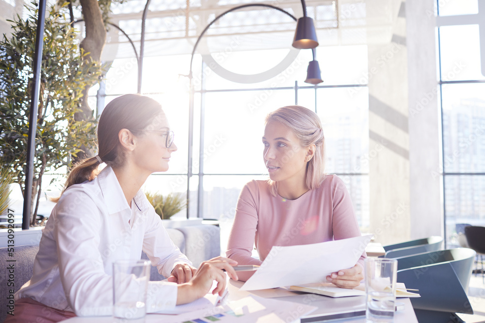 Thoughtful young businesswomen in formalwear sitting at table in cafe full of light and examining important papers Thoughtful young businesswomen in formalwear sitting at table in cafe full of light and examining important papers