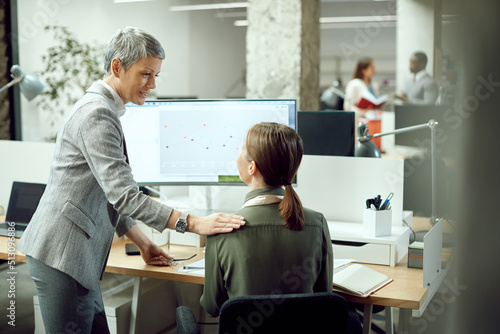 Happy female CEO talks to businesswoman who is working on desktop PC in office.