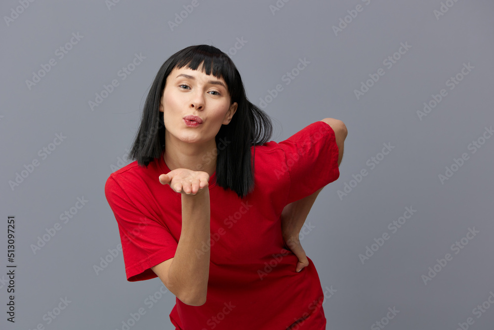 a sweet, pleasant, beautiful woman in a red T-shirt stands on a gray background and sends an air kiss to the camera smiling pleasantly a sweet, pleasant, beautiful woman in a red T-shirt stands on a gray background and sends an air kiss to the camera smiling pleasantly