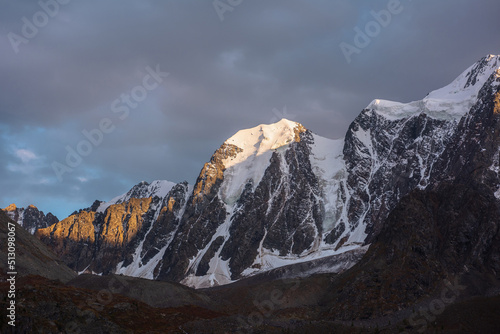 Gloomy landscape with giant snowy mountain top and sunlit gold rocks in dramatic gray sky. Huge snow mountains with hanging glacier and cornice under rainy clouds. Snow-covered mountains in overcast.