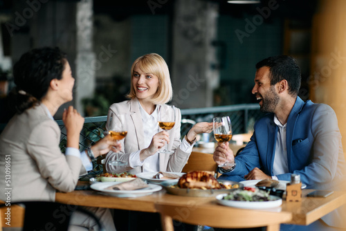 Cheerful businesswoman and her coworkers drink wine and talk during lunch at restaurant.