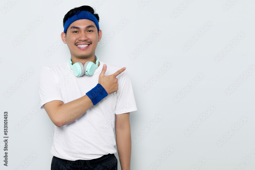 Portrait of smiling young Asian sportman in blue headband and sportswear white t-shirt with headphones, pointing fingers at copy space, advertisement banner isolated on white background Portrait of smiling young Asian sportman in blue headband and sportswear white t-shirt with headphones, pointing fingers at copy space, advertisement banner isolated on white background