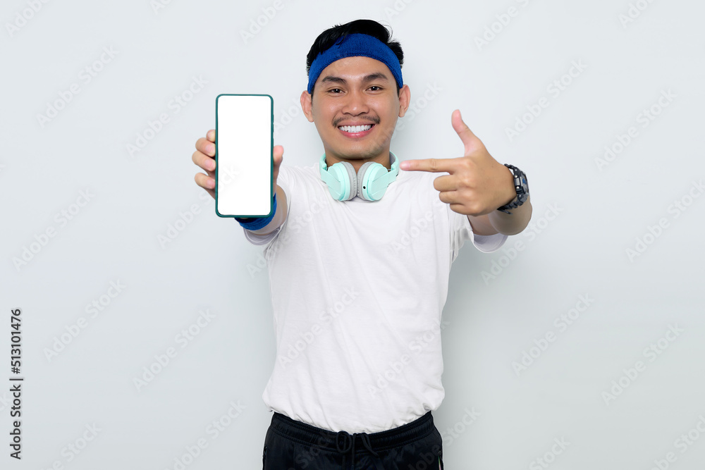 Portrait of Smiling young Asian sporty man in blue headband and white t-shirt with headphones, showing mobile phone blank screen recommending app isolated on white background. Workout sport concept Portrait of Smiling young Asian sporty man in blue headband and white t-shirt with headphones, showing mobile phone blank screen recommending app isolated on white background. Workout sport concept