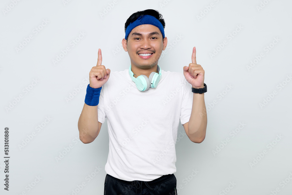 Fototapeta premium Excited young Asian sportman in blue headband and sportswear white t-shirt with headphones, pointing fingers up having a good idea isolated on white background. Workout sport concept