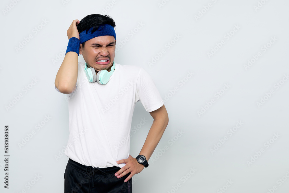 Stressed young Asian sportman in blue headband and sportswear white t-shirt with headphones, touching his temples and having headache isolated on white background. Workout sport concept Stressed young Asian sportman in blue headband and sportswear white t-shirt with headphones, touching his temples and having headache isolated on white background. Workout sport concept
