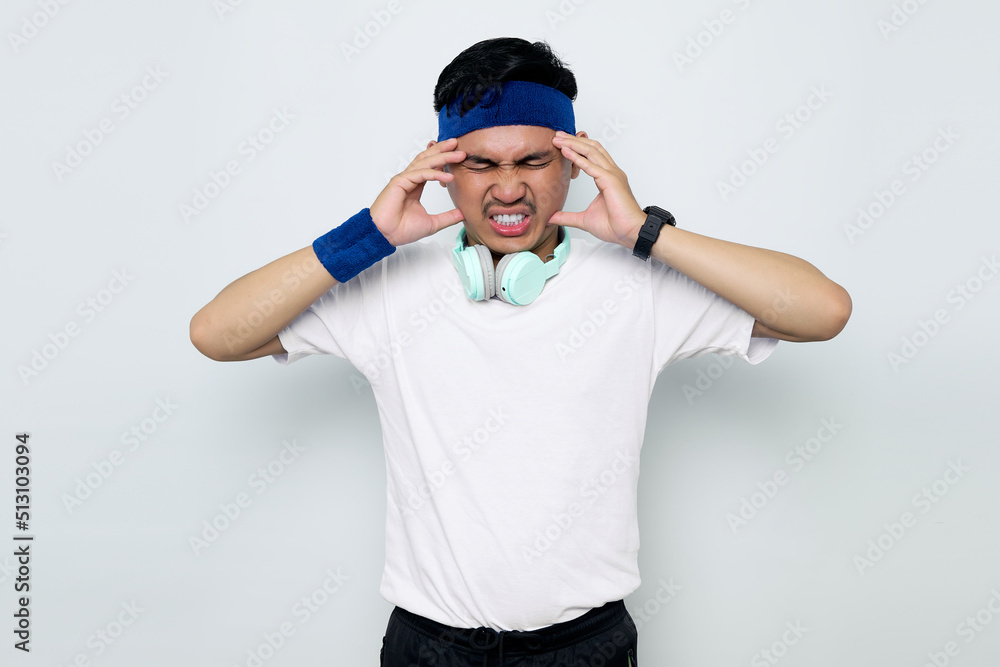 Fototapeta premium Stressed young Asian sportman in blue headband and sportswear white t-shirt with headphones, touching his temples and having headache isolated on white background. Workout sport concept