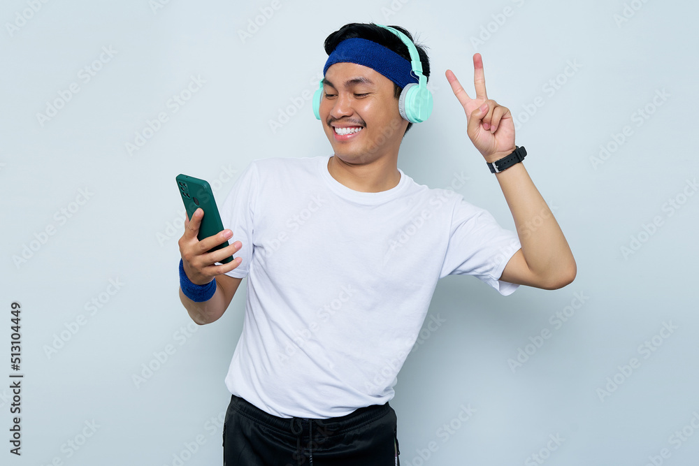 smiling young Asian sportman in blue headband and sportswear white t-shirt while listening music favorite with headphones, doing selfie shot on mobile phone, peace gesture isolated on white background smiling young Asian sportman in blue headband and sportswear white t-shirt while listening music favorite with headphones, doing selfie shot on mobile phone, peace gesture isolated on white background