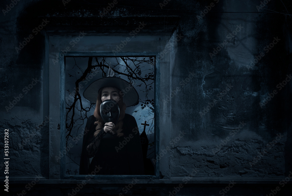 Halloween witch holding black face mask standing in old damaged window with wall over cross, church, birds, dead tree and spooky cloudy sky, Halloween mystery concept
