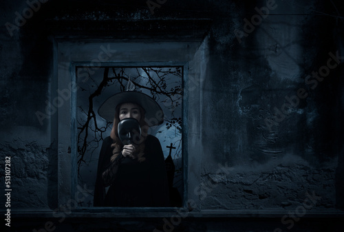 Halloween witch holding black face mask standing in old damaged window with wall over cross, church, birds, dead tree and spooky cloudy sky, Halloween mystery concept