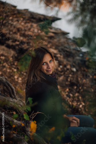 Young Caucasian woman sitting on tree roots in forest, smiling, looking at camera and posing near river with tree in background, wearing blue jeans and black jacket. Autumn walk in forest.
