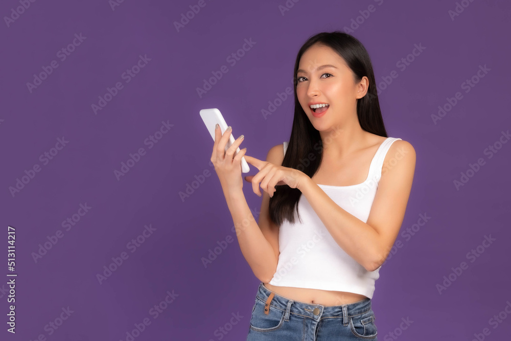 Beautiful Asian brunette woman cute girl in white tank top playing a smart phone isolated on purple background - mobile phone uses a online banking for pay online shopping Beautiful Asian brunette woman cute girl in white tank top playing a smart phone isolated on purple background - mobile phone uses a online banking for pay online shopping