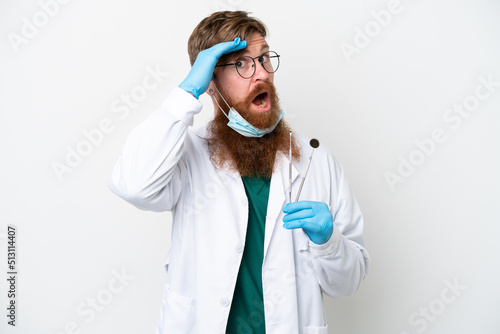 Dentist reddish man holding tools isolated on white background doing surprise gesture while looking to the side