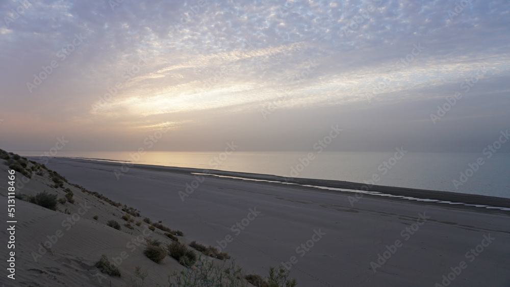 Dawn on the sandy beach of the sea. Sand dune. The sun's rays peek out from behind the clouds. Car tracks in the sand. A light wave, almost calm. The water merges with the sky. Autumn morning